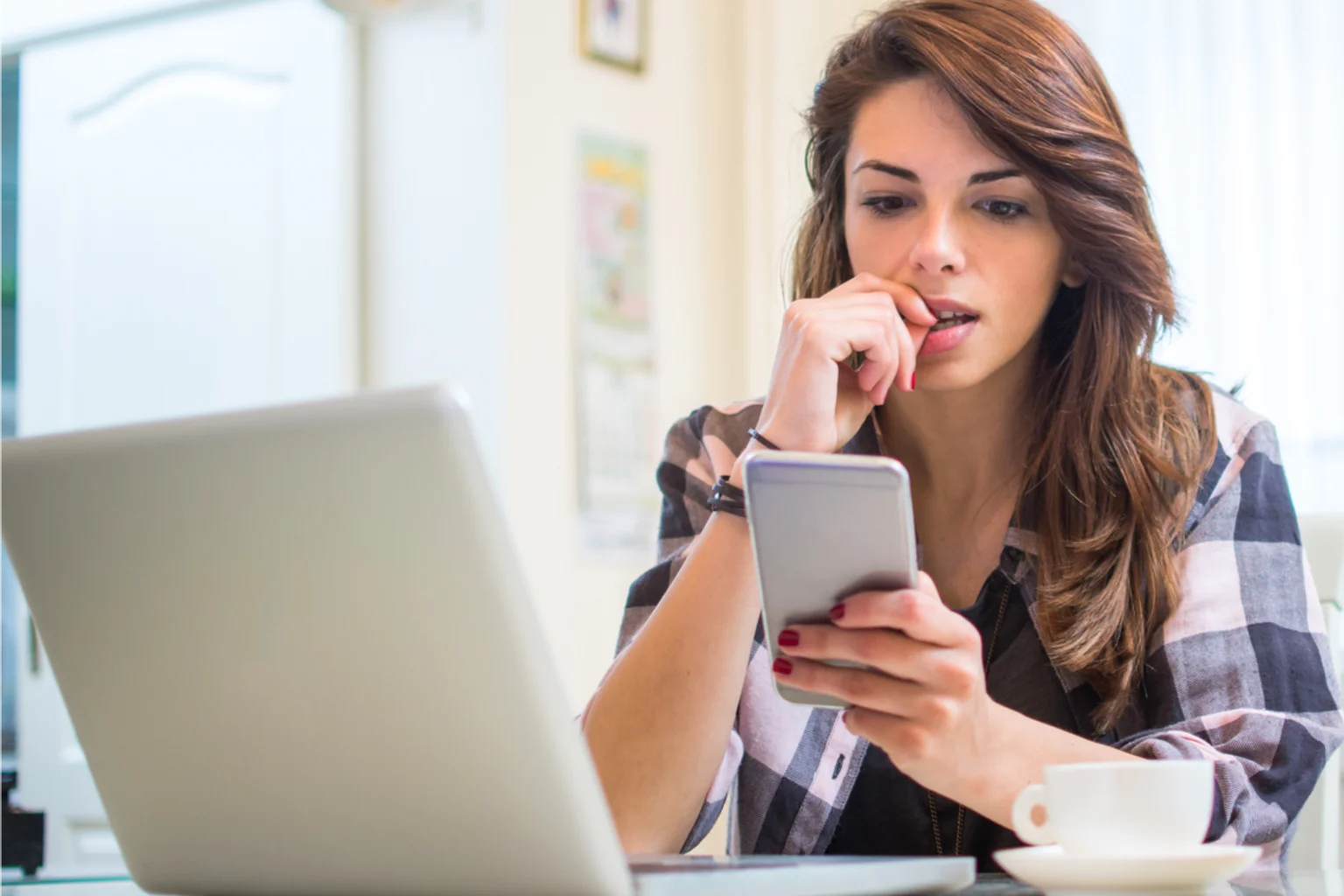 A woman looks at her phone beside a laptop and a cup of coffee.