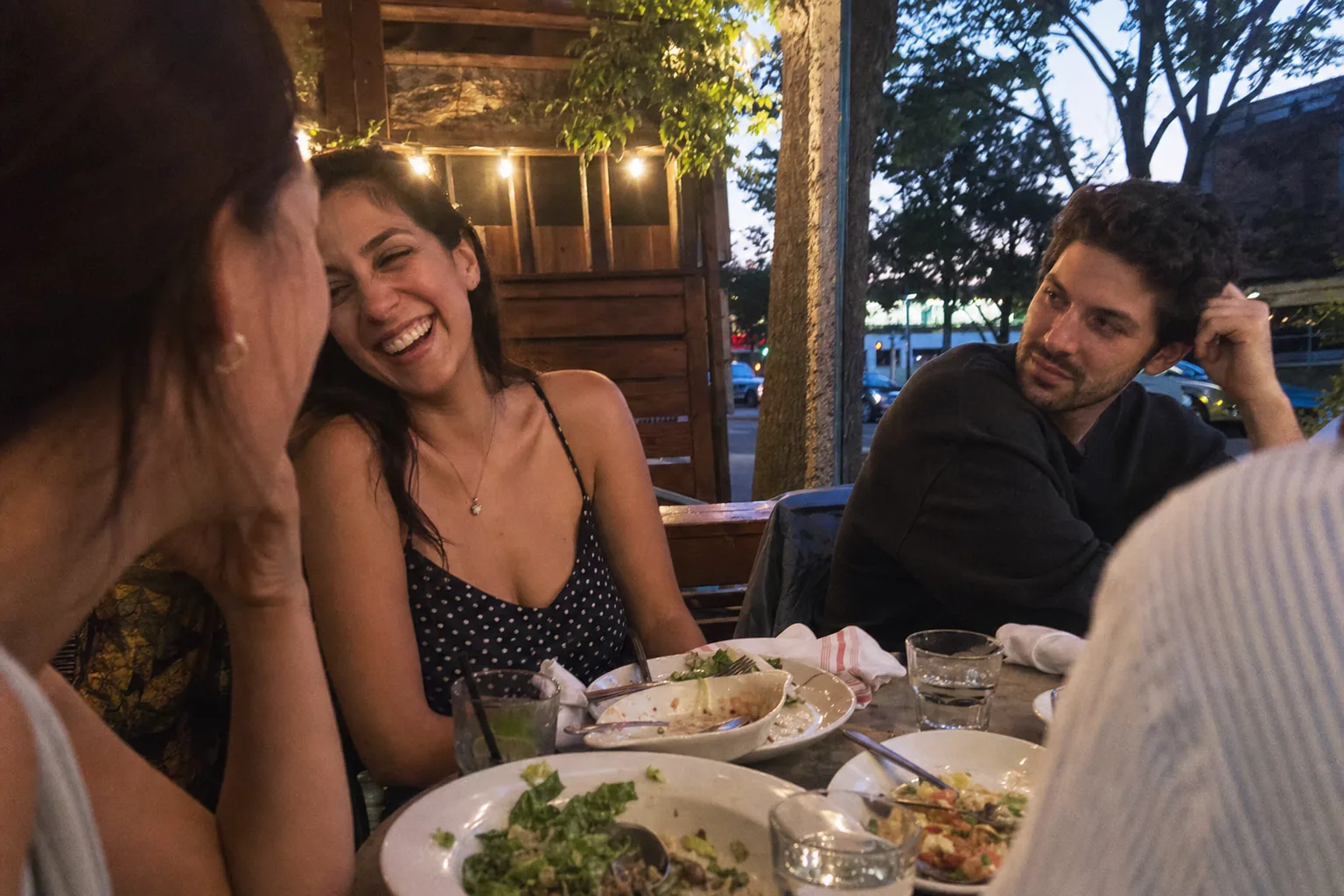 A woman laughs with friends while a man across the dinner table looks toward her with quiet interest.