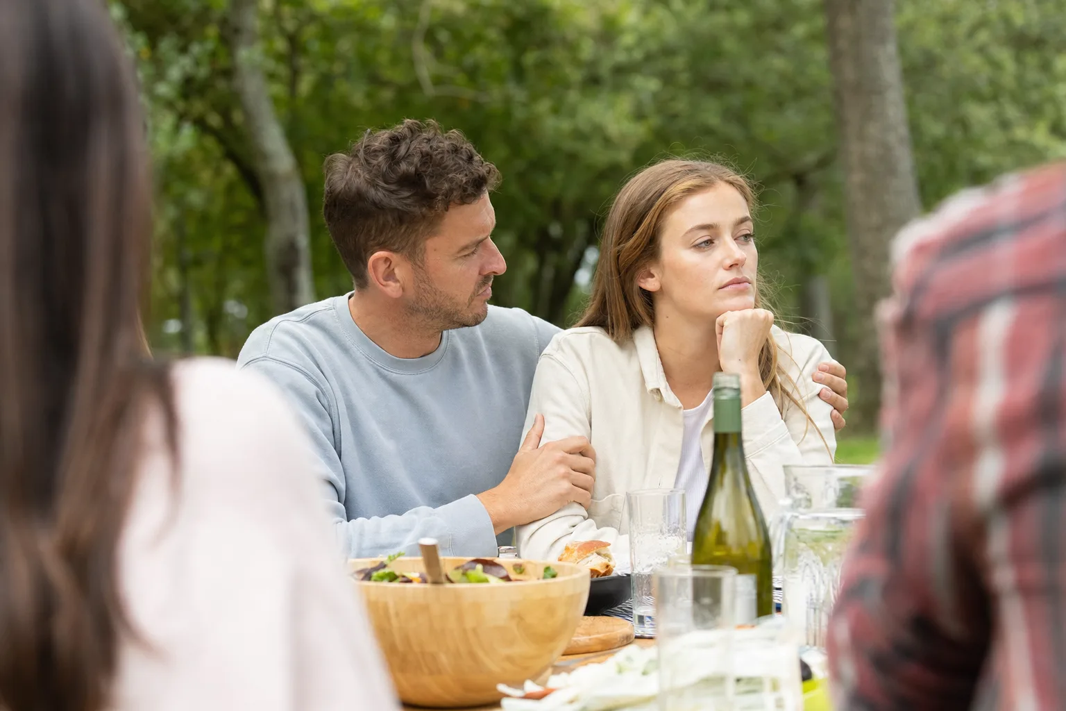 A couple sits together at a group picnic while one partner looks quietly distant.