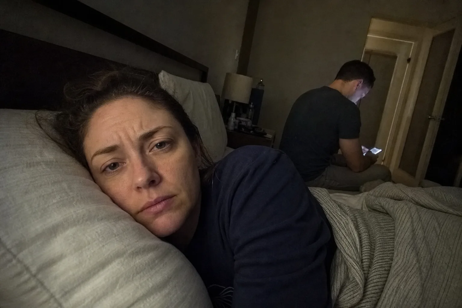 A worried person lies awake while their partner texts at the edge of the bed.