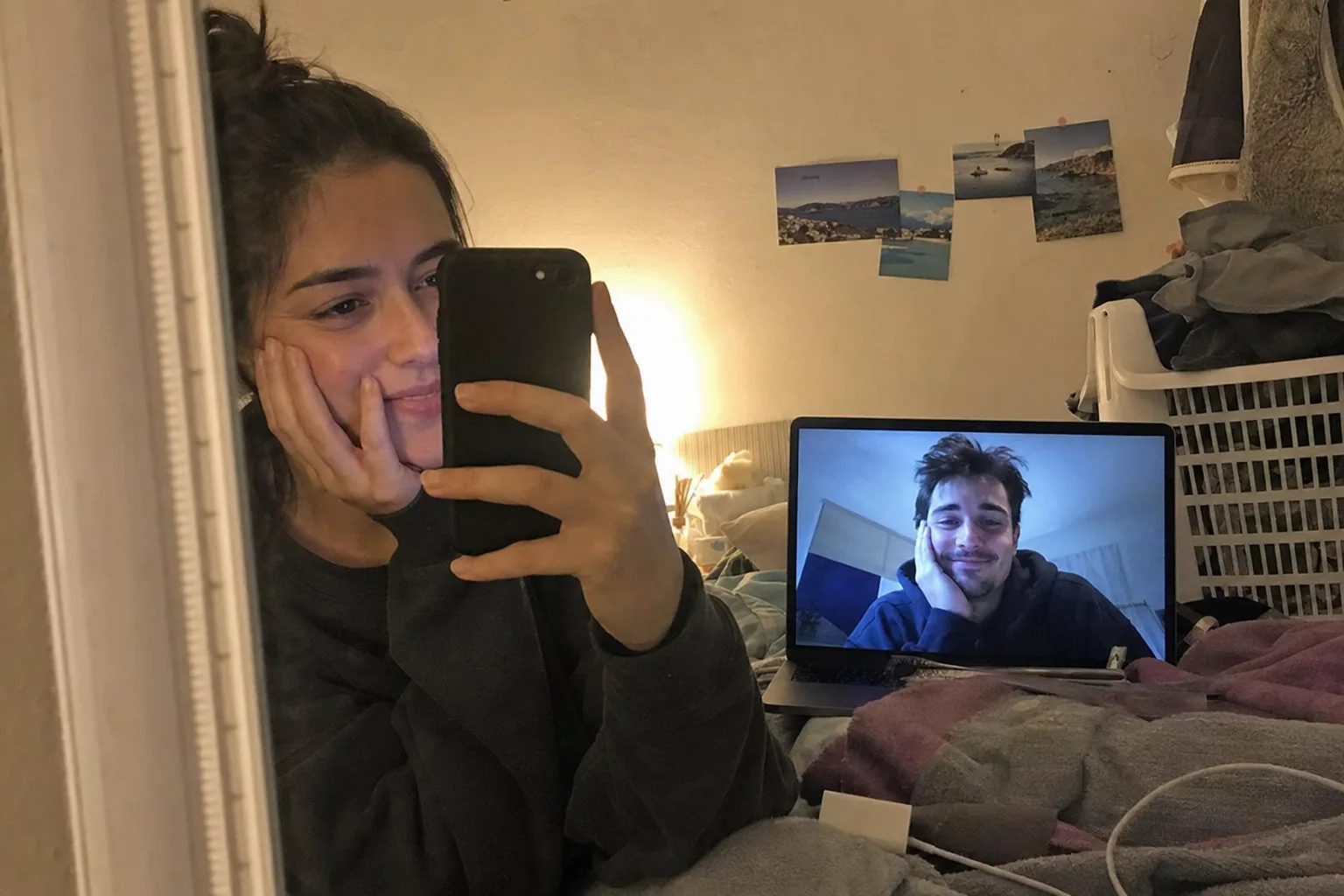 A woman takes a bedroom mirror selfie beside a laptop video call with her long-distance partner.