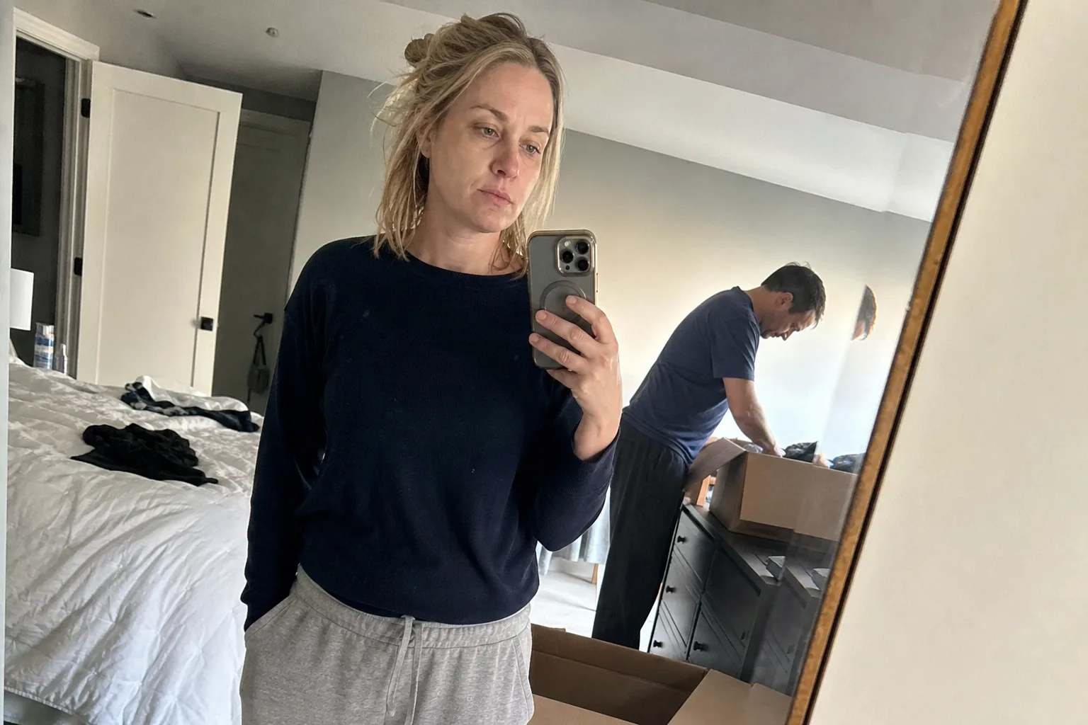 A woman takes a mirror selfie while her spouse packs in a half-empty bedroom.